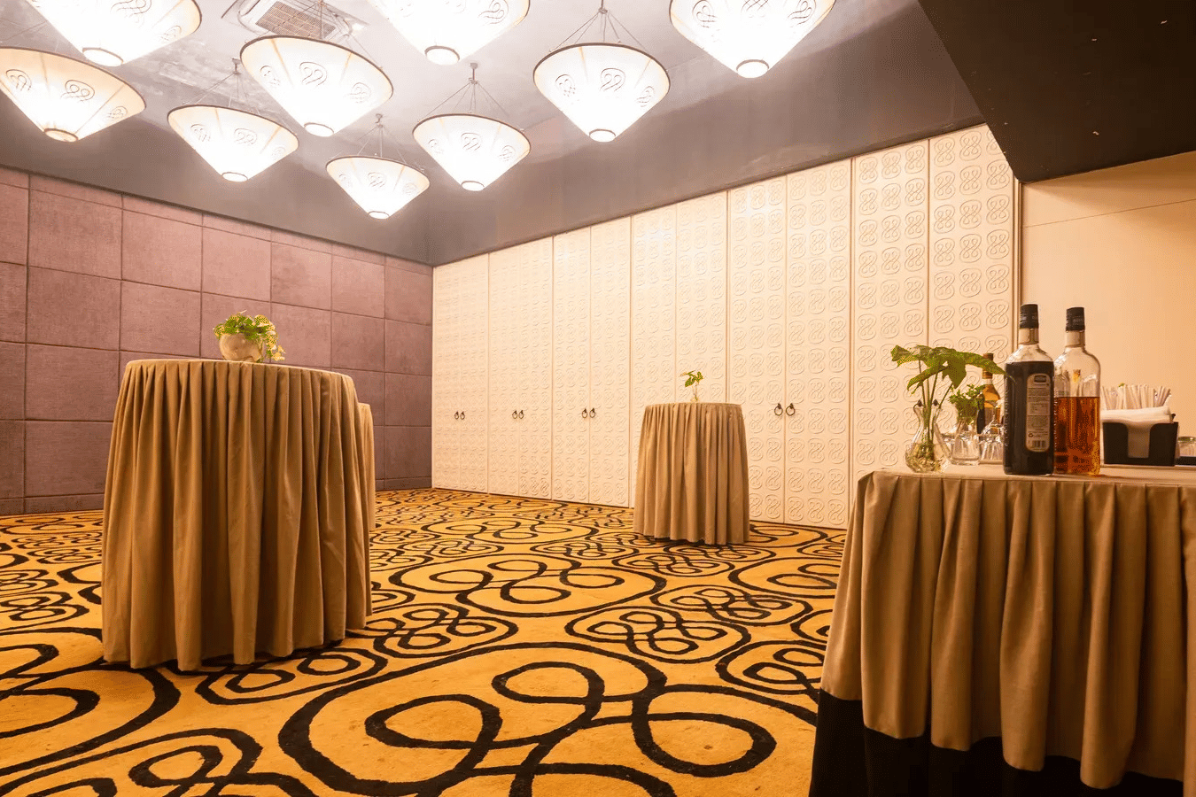 Standing cocktail tables with beige covers in Dholavira banquet hall, featuring patterned carpets, hanging inverted cone lights, and a bar counter at 3102bce - A Vedic Resort by Lindsay.