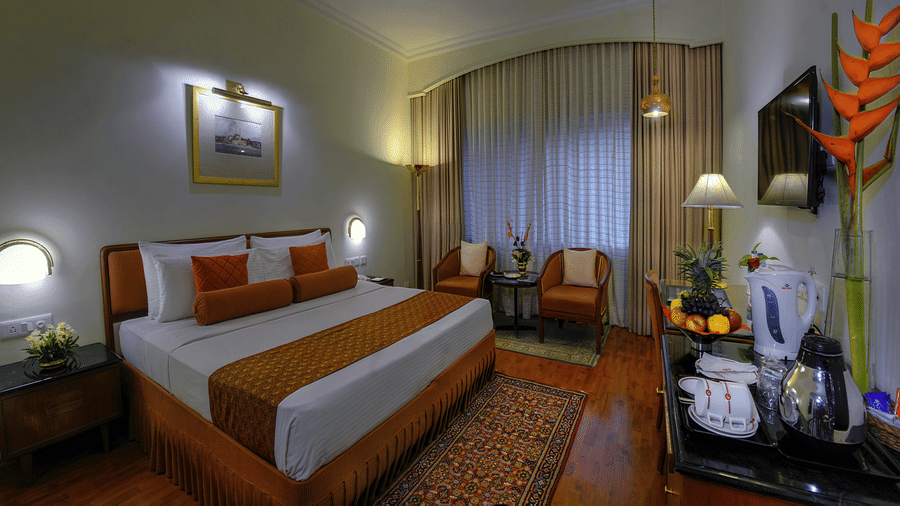 A neatly-made bed facing a wall-mounted television and a seating area in a Premier Room at The Clarks Varanasi. 
