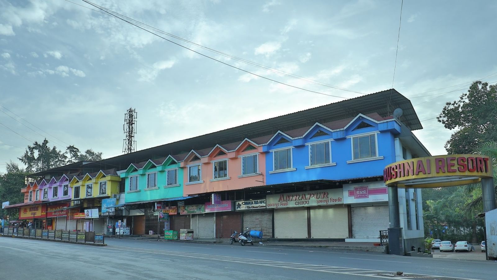 The Krushnai Resort sign marks a long building with colourful facade lining a quiet road under a cloudy sky.