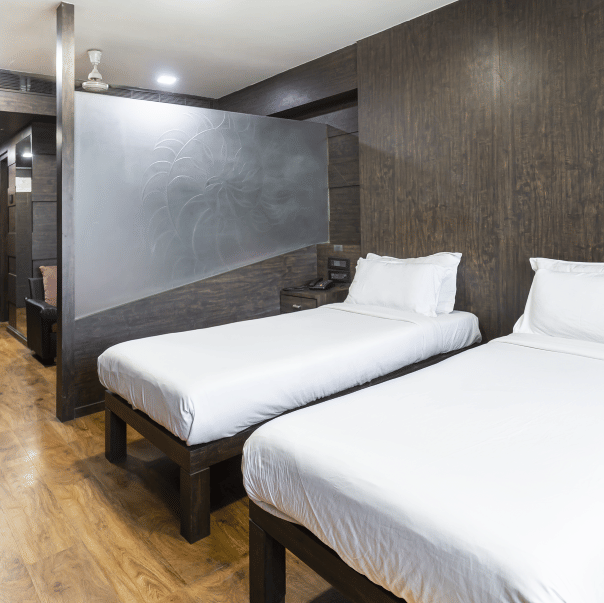 A room with two single beds, dark textured walls, a ceiling light, and a compact open wardrobe unit at Infiniti Hotel, Indore.