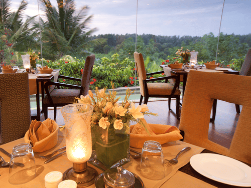 Candle-lit dining tables with plates, cutlery, napkins and floral centrepieces placed indoors, facing large glass windows that overlook greenery and palm trees at the best restaurant in Chikmagalur inside The Serai Chikmagalur.