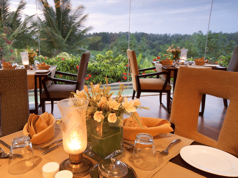 Candle-lit dining tables with plates, cutlery, napkins and floral centrepieces placed indoors, facing large glass windows that overlook greenery and palm trees at the best restaurant in Chikmagalur inside The Serai Chikmagalur.