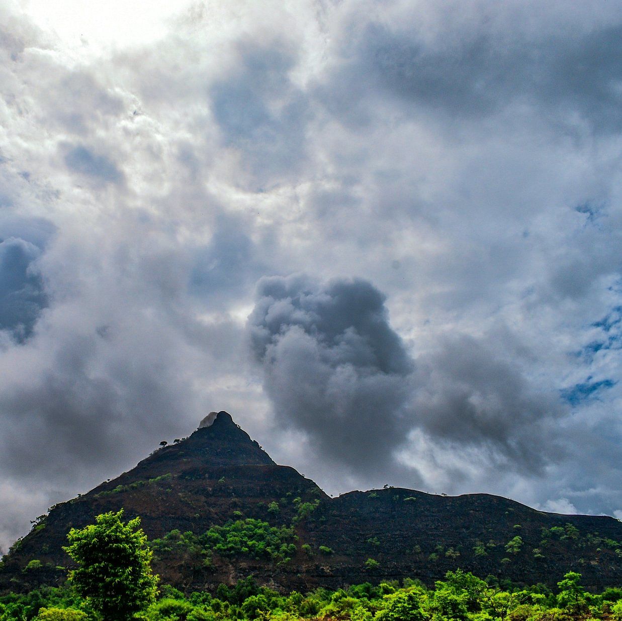 A dramatic sky filled with storm clouds over a conical mountain peak, surrounded by green foliage.