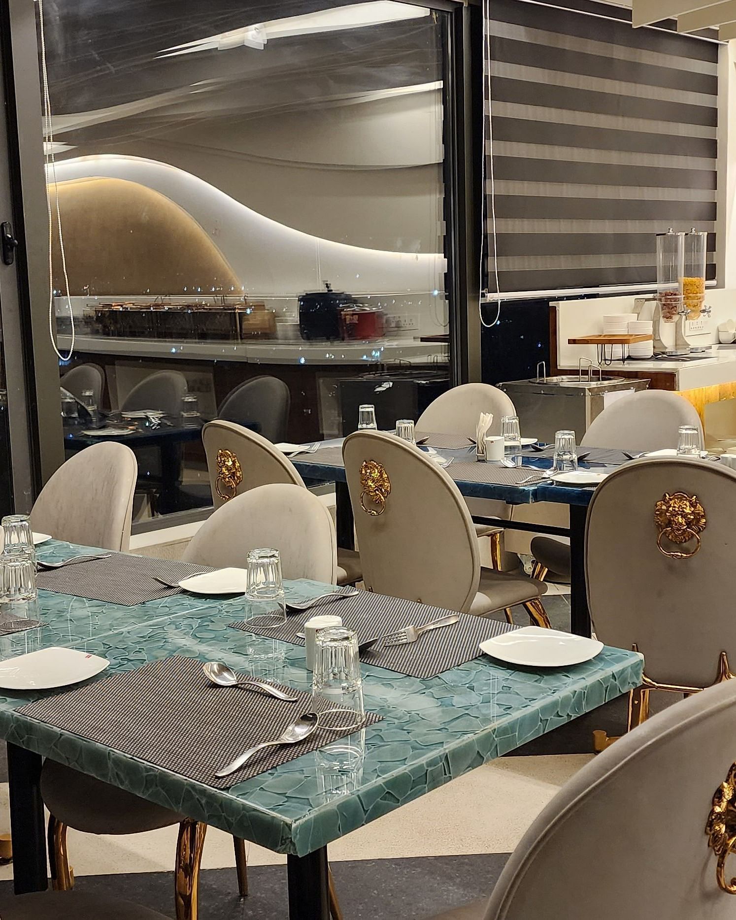 An elegant dining area with white and gold chairs and set tables, located beside large windows offering a view outside.