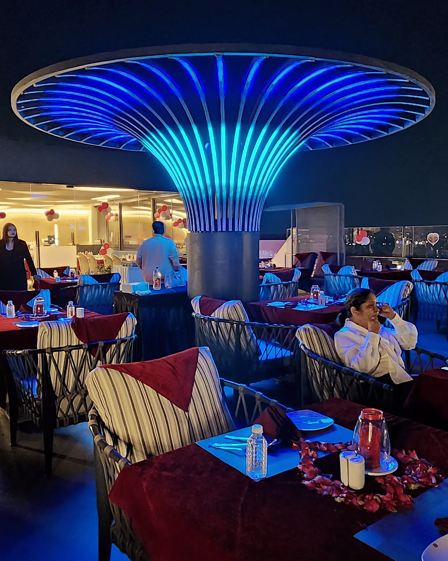 A vibrant restaurant interior with modern blue lighting and red chairs, featuring a central decorative light fixture shaped like a mushroom.