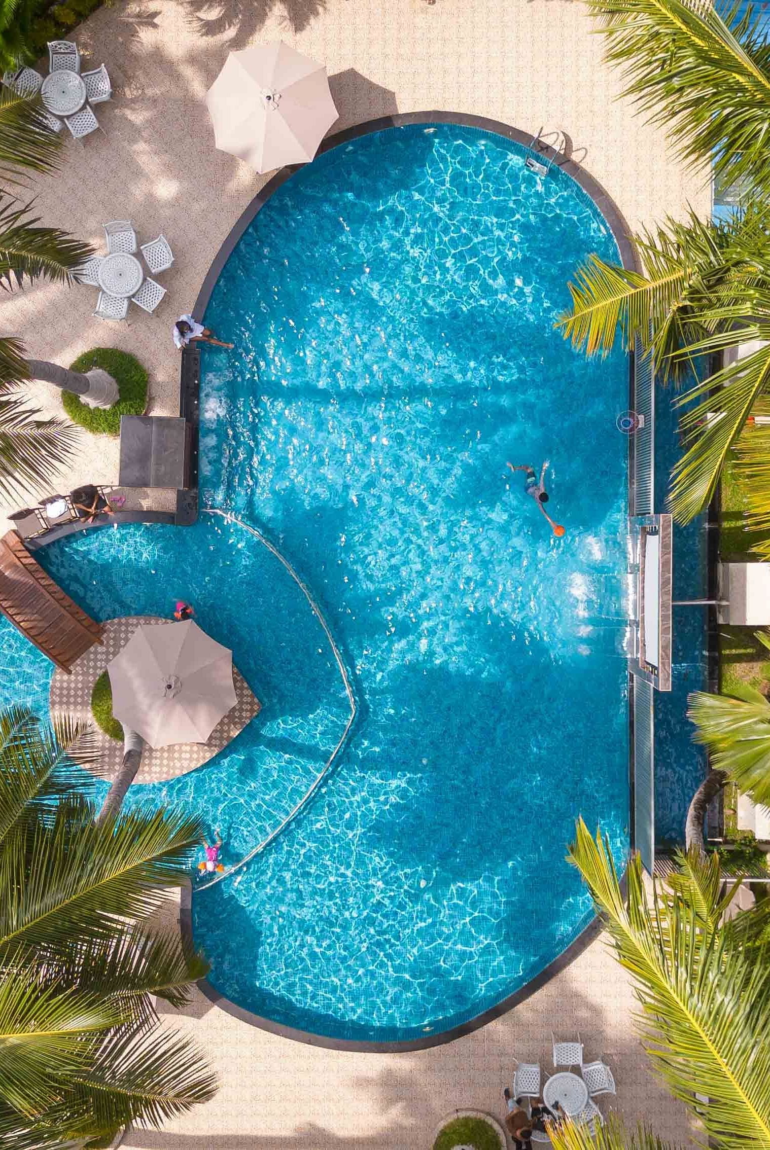 An aerial view of a stunning pool surrounded by palm trees and lounge chairs, with a peaceful, tropical environment surrounding the area. The pool is the centre of this serene outdoor setting - Coral Reef Resort & Spa, Havelock
