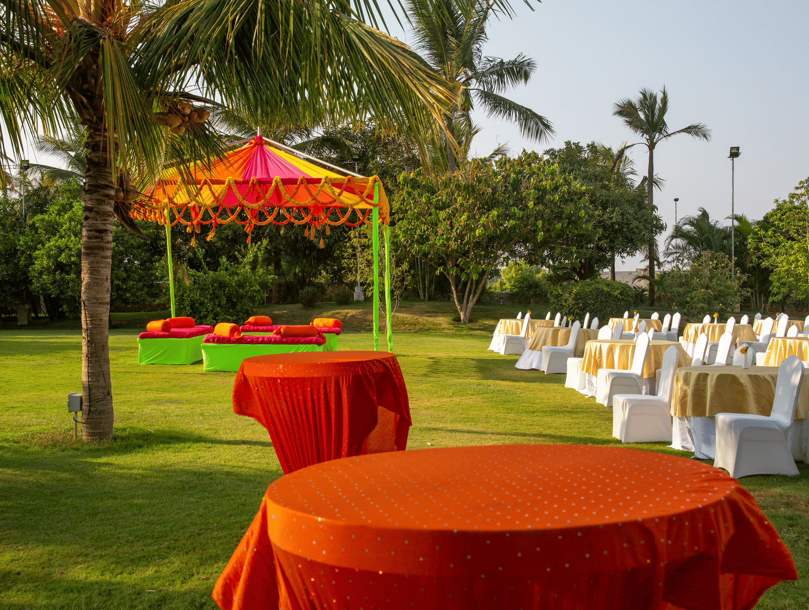 An outdoor dining area with orange-draped tables on a green lawn with palm trees and a vibrant tent - Grande Bay Resort & Spa, Mamallapuram