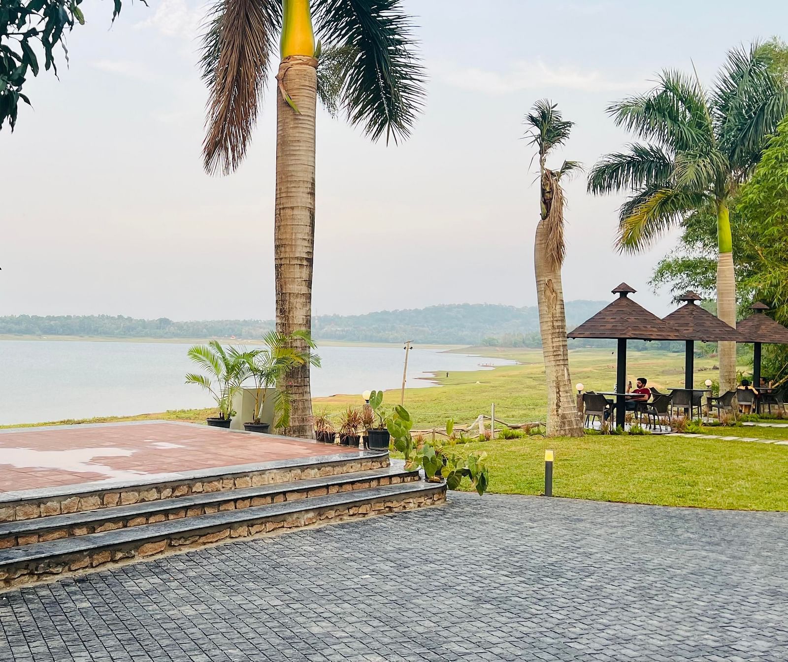 View of a lakeside area with stone steps, palm trees, and a paved pathway. A gazebo with a thatched roof sits near the water, with grassy areas and distant hills visible across the lake