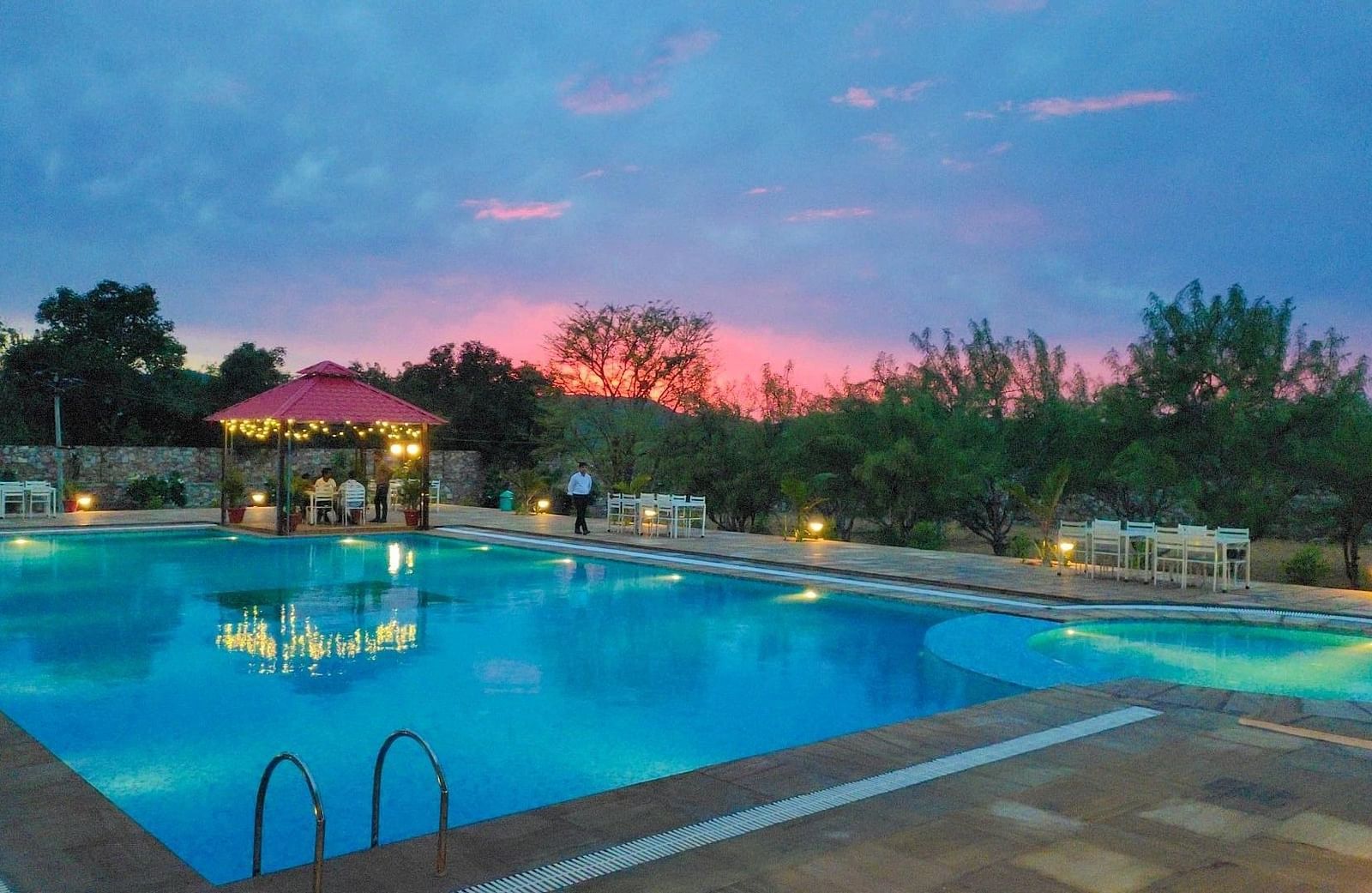 An outdoor swimming pool at dusk with a pink and blue sky in the background - Bamboo Saa Mulberry Resort, Pushkar