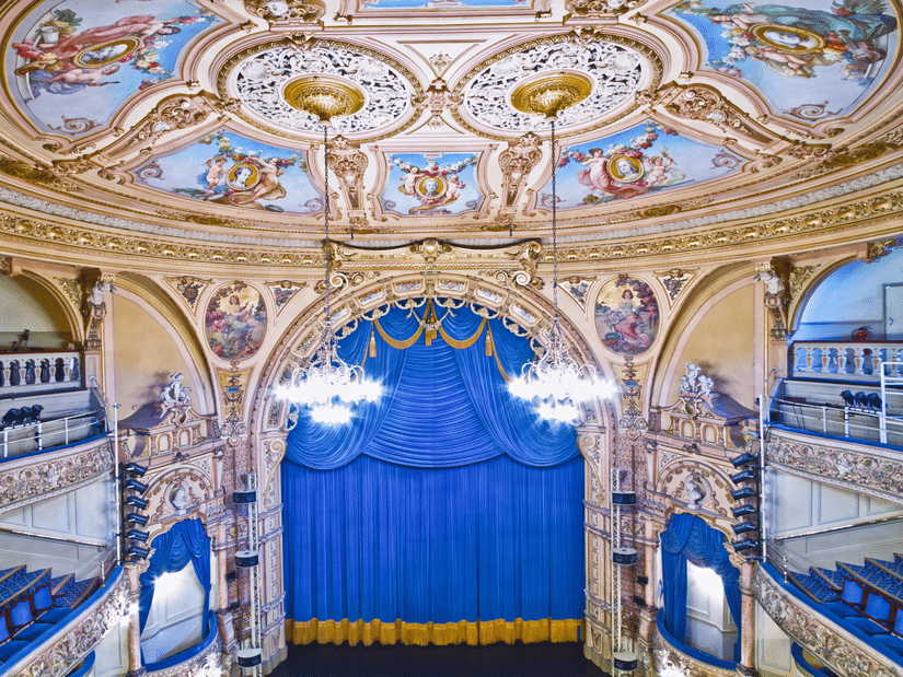 A view from above of a theatre's stage with blue curtains covering it and art on the roof and chandeliers hanging from it.