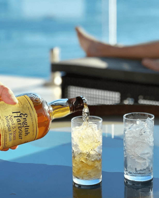 A hand pours a drink from a bottle into a glass filled with ice, set against a blurry background of water.