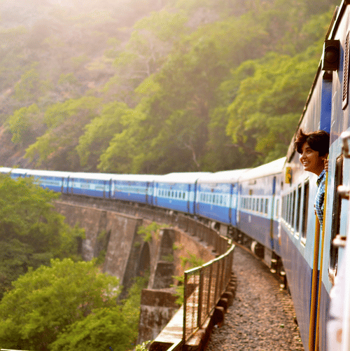 A view of Bangalore to Kolkata train travelling on a bridge next to a mountain with many trees and a person looking at the nature from the door.