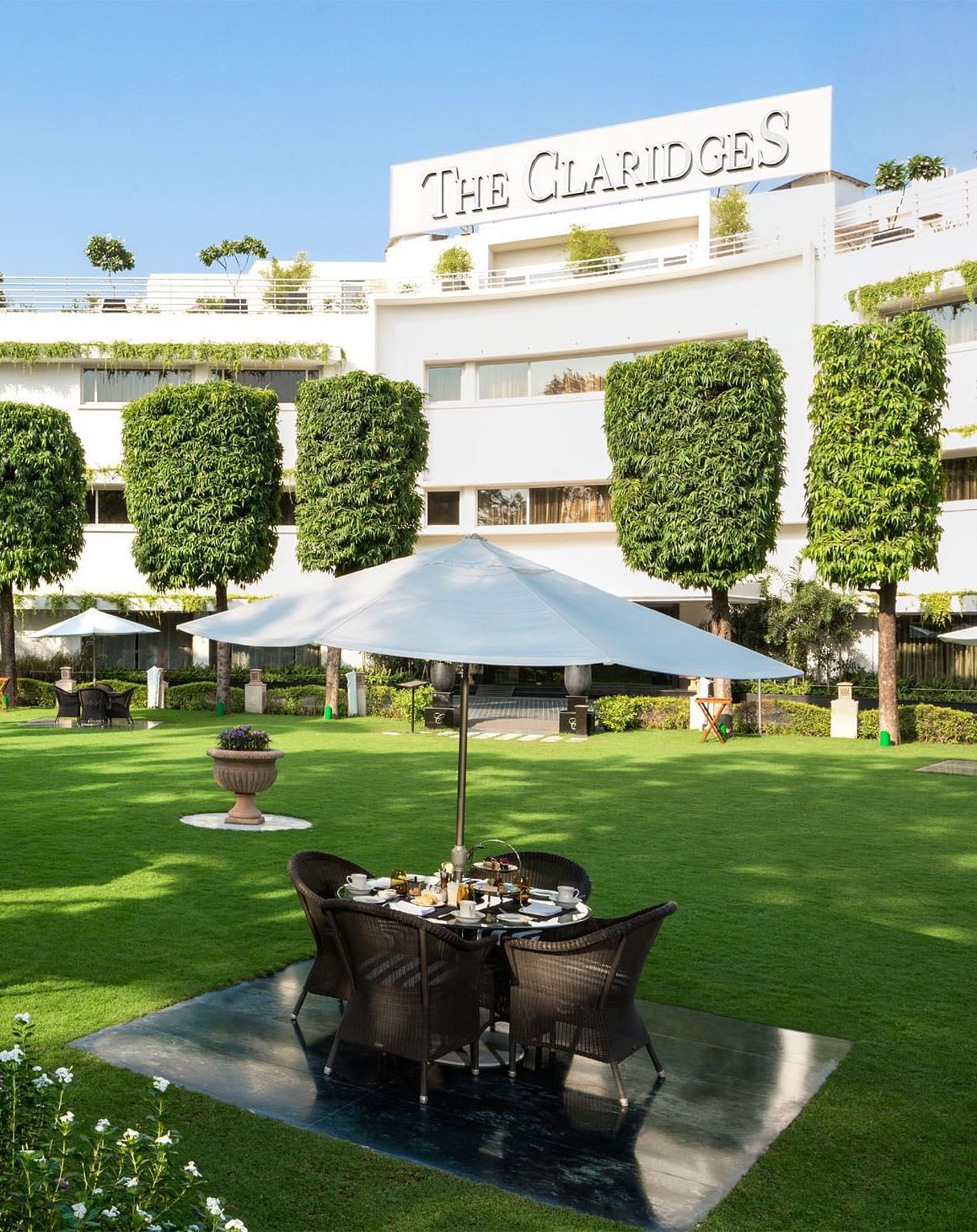 Facade of The Claridges New Delhi, featuring a green garden in the front and surrounded by towering trees, offering a serene and elegant ambience