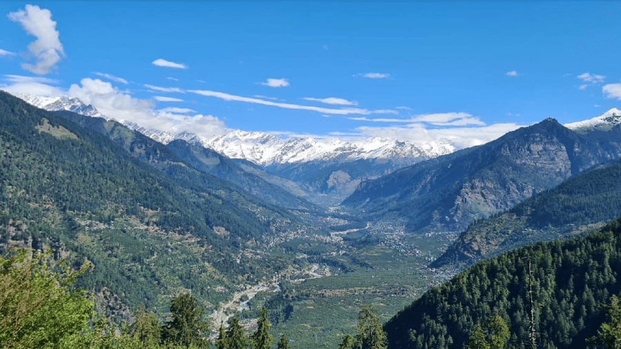 A panoramic view of the valley with towering, lush green mountains in the foreground and majestic snow-capped Himalayan peaks under a bright blue, cloudy sky seen from Amara Upepo - The Sky Village, Manali.
