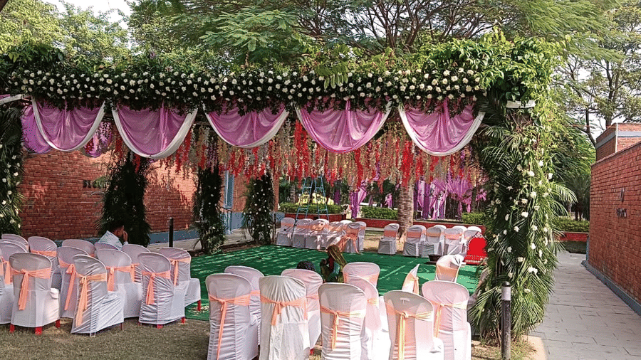A decorated outdoor seating area for an event, featuring white chairs with pink sashes and a canopy covered in green leaves and pink flowers | Nandan Resort