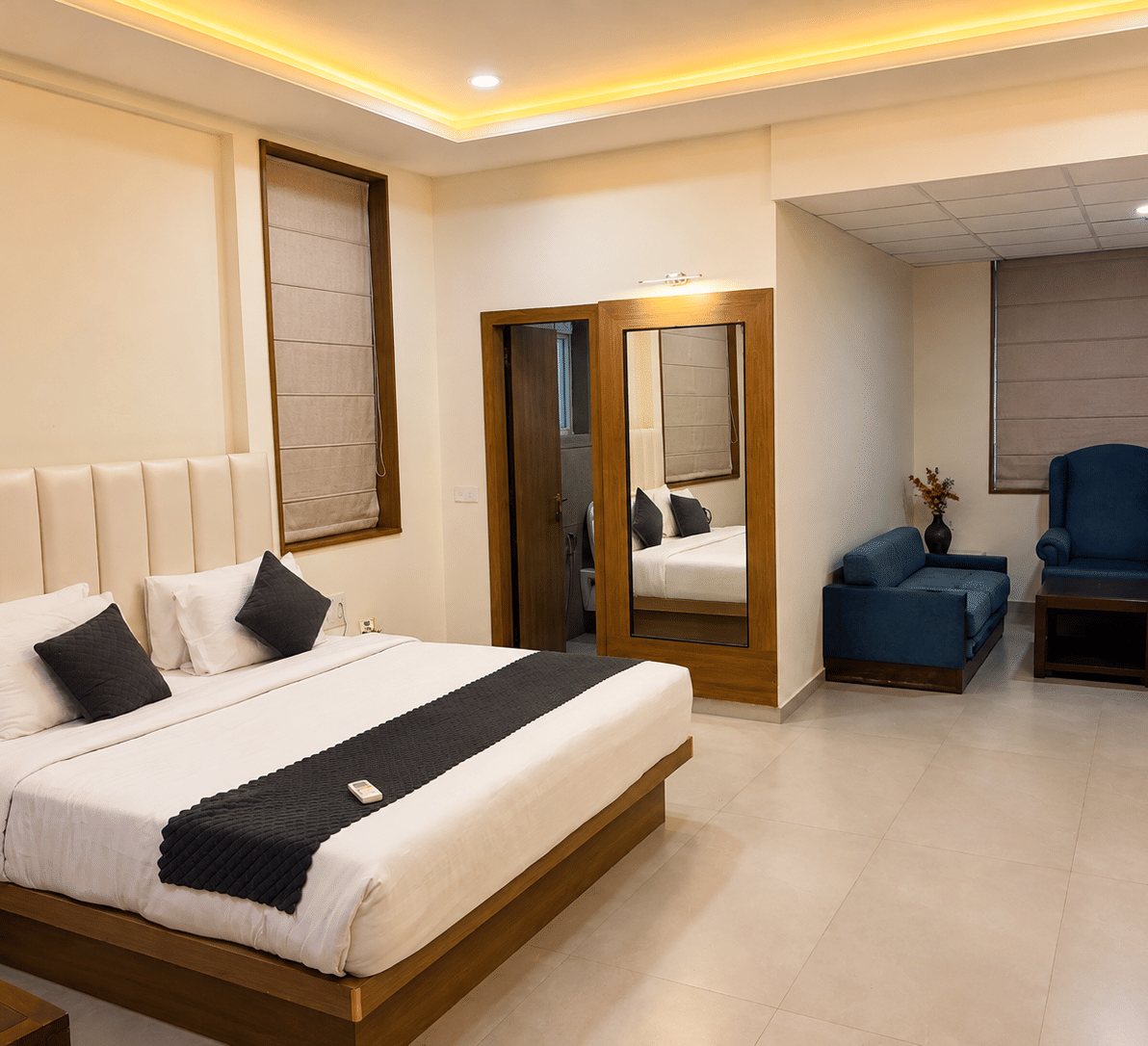 Suite at TGI Apple Inn, Ajmer Road, Jaipur featuring a bed with pillows, a seating area with armchairs and a table, blinds on the windows, and a doorway.