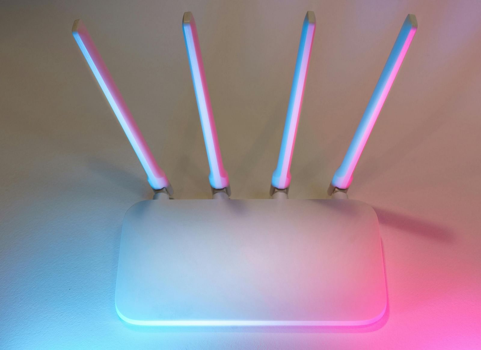 A white wireless router with four antennae illuminated in blue and pink hues against a neutral wall.