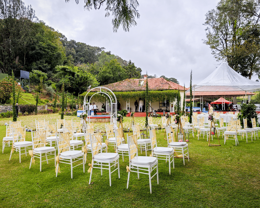 n outdoor event setting with a floral archway and a seating area with white chairs and yellow sashes on a green lawn, surrounded by lush foliage and trees.