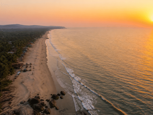 Aerial view of Mandrem Beach at sunset with palm-lined sand, thatched beach huts, waves rolling in, and the sun dipping toward the Arabian Sea.