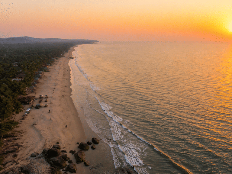Aerial view of Mandrem Beach at sunset with palm-lined sand, thatched beach huts, waves rolling in, and the sun dipping toward the Arabian Sea.