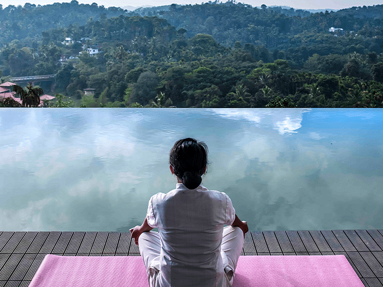 Person meditating beside an infinity pool overlooking lush green hills.