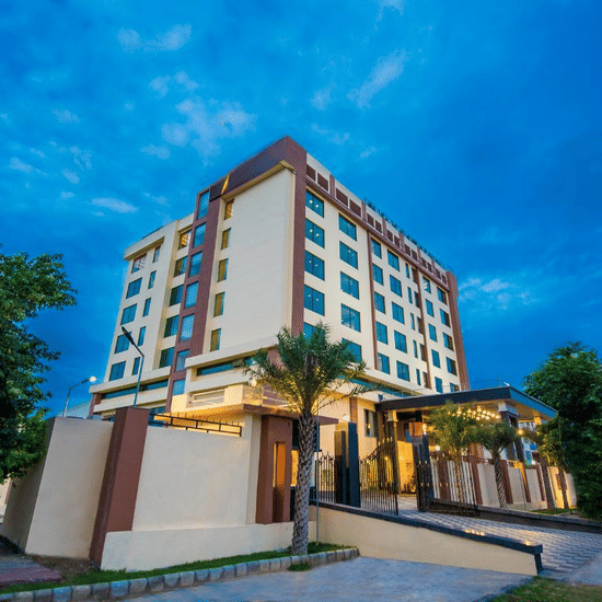 An angled evening view of a contemporary hotel building illuminated against a twilight sky, featuring a paved driveway and greenery at Golden Tulip Kukas, Jaipur.