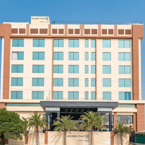 A modern, multi-storey hotel with a beige facade and palm trees at the entrance. Bright, clear daylight with a blue sky background at Golden Tulip Kukas, Jaipur.