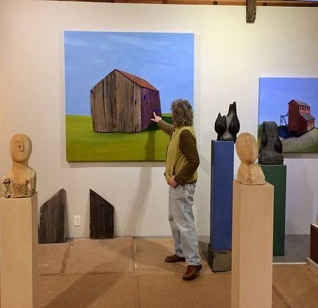 A person in an art studio pointing at a painting of a rustic barn, surrounded by sculptures and other artworks.