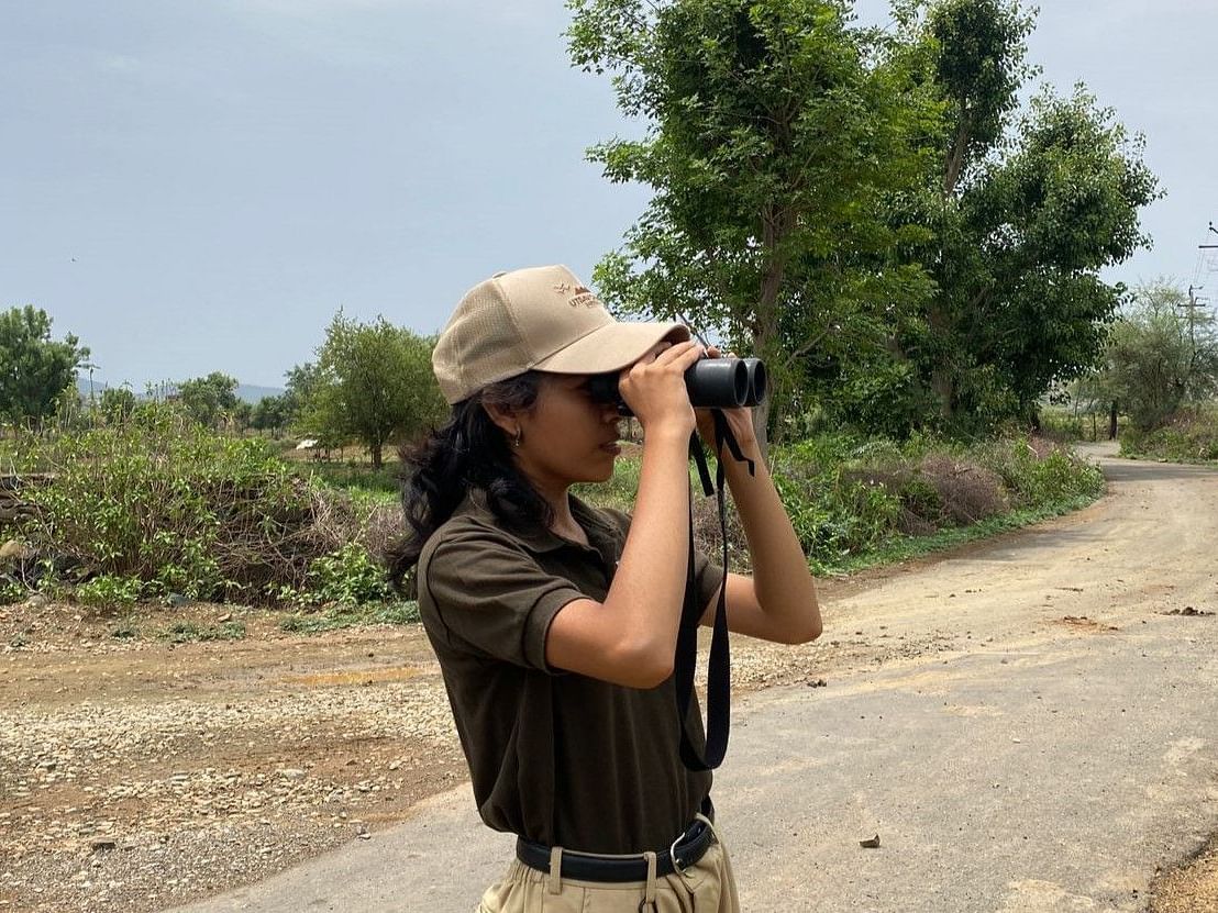 Woman in safari uniform using binoculars on a rural path with trees in the background
