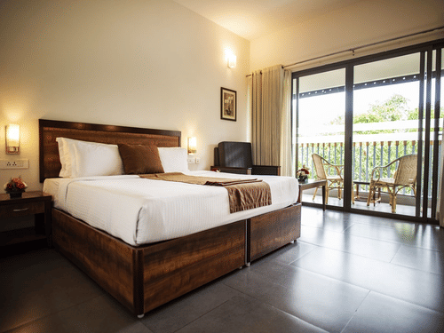 Bright bedroom at Abad Green Forest, Thekkady with wooden furnishings, crisp white bedding, and sliding doors opening to a balcony overlooking greenery.