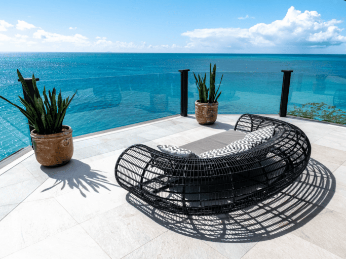An outdoor seating area with a modern chair and potted plants overlooks a vibrant turquoise ocean under a bright blue sky.