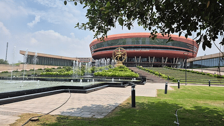 Exterior view of a modern circular convention centre building featuring glass facade design, surrounding garden space, paved pathway in front.