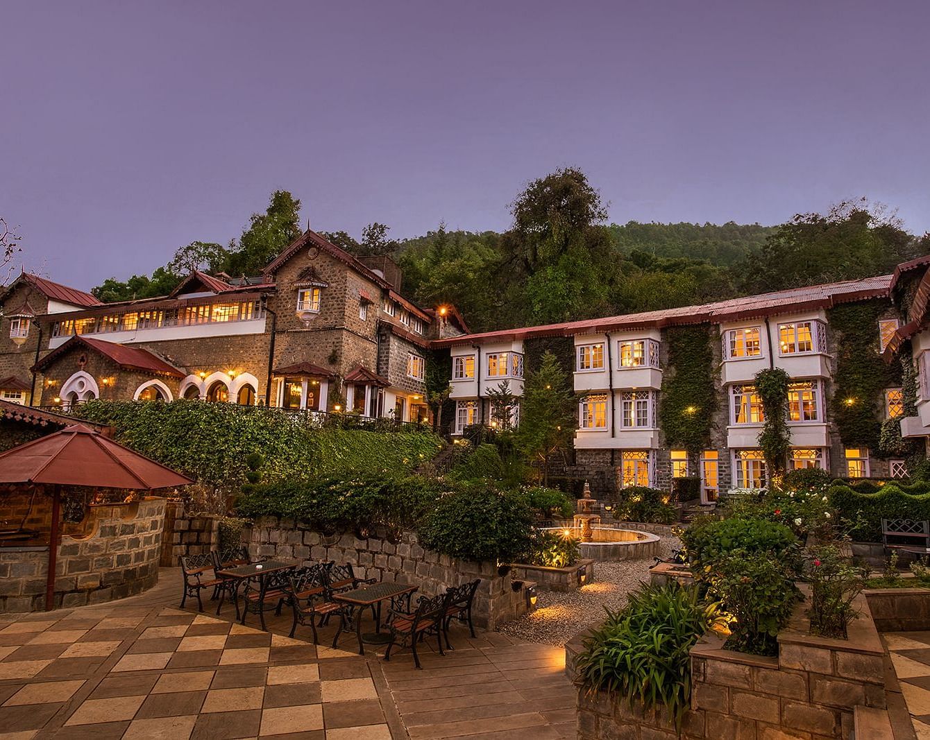 A scenic view of a the naini retreat, nainital, The resort features multiple buildings with stone exteriors and white trim. The surrounding landscape includes mountains, trees, and a winding pathway.