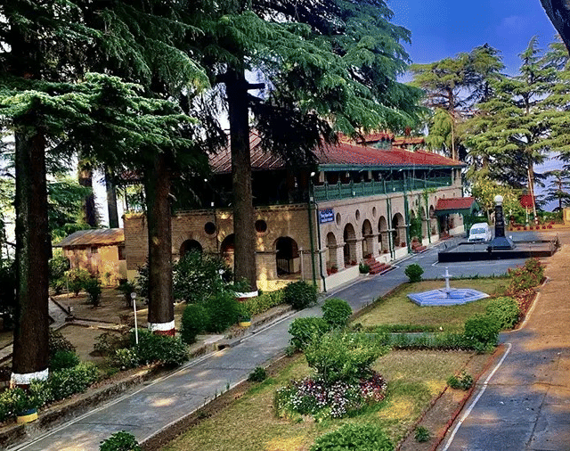  An aerial-style view of a colonial-era building surrounded by a neat garden with a fountain, bordered by a paved path and tall trees.