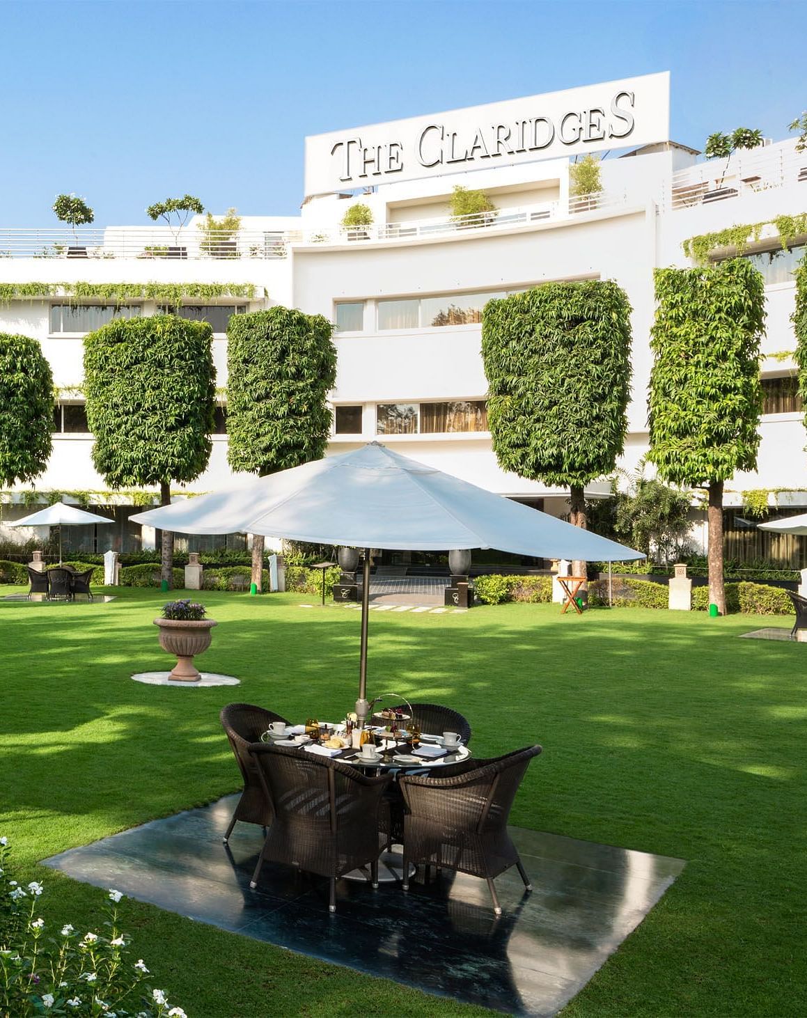 Facade of The Claridges New Delhi, featuring a green garden in the front and surrounded by towering trees, offering a serene and elegant ambience
