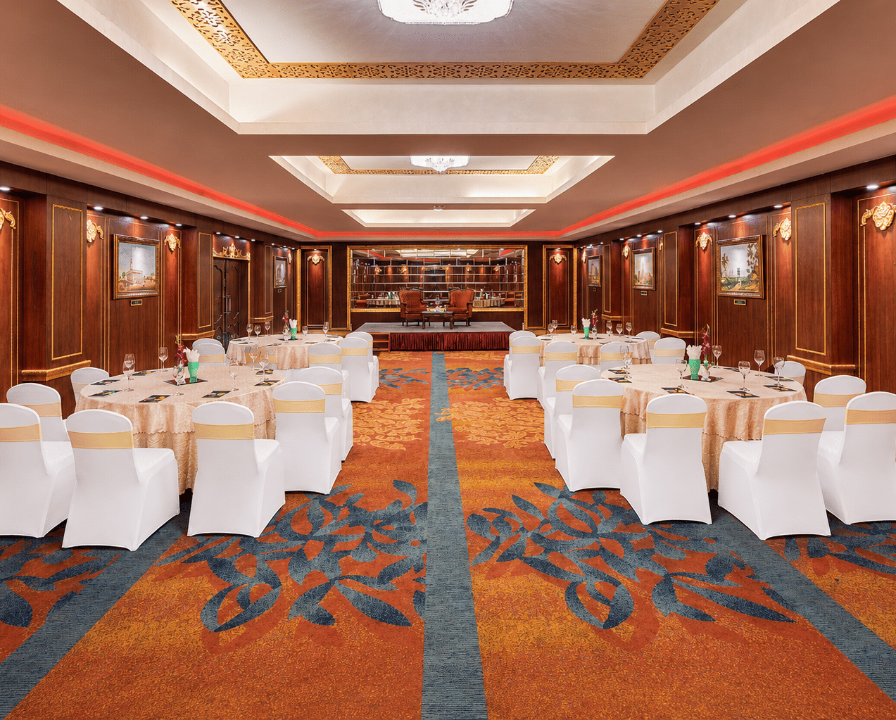Tulip banquet hall with round tables, white chair covers, patterned carpet, wood-panelled walls with framed paintings and a stage at MAYFAIR Garden, Rourkela.