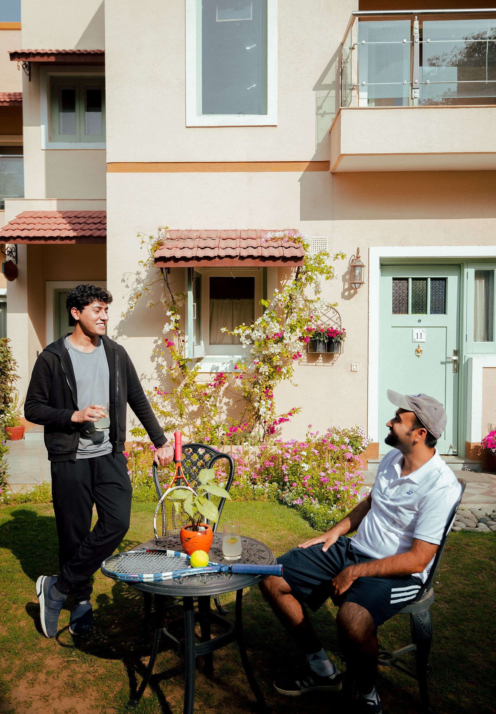 Two men near a round table with drinks in a garden outside a cream-coloured building with balconies and flowering plants | Karma Chalets