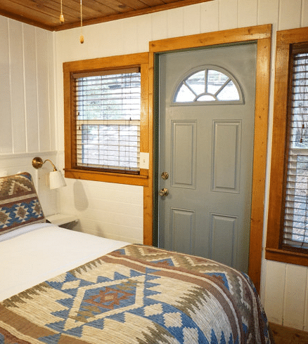 Overview of a Room at Shaver Lake Village Hotel with a bed featuring a patterned blanket, a grey door with a half-circle window, and two windows with blinds.