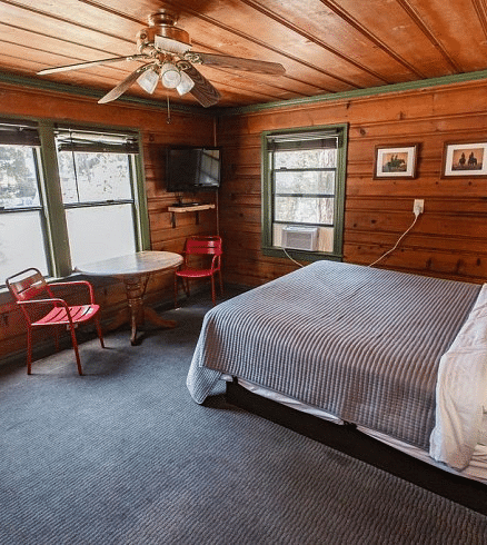 a Cozy bedroom at Shaver Lake Village Hotel with wood paneled walls, two windows, a double bed, and red chairs next to a small table.