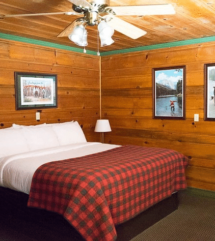 a Bedroom at Shaver Lake Village Hotel with wooden walls, plaid comforter, white pillows, framed artwork, and a ceiling fan.
