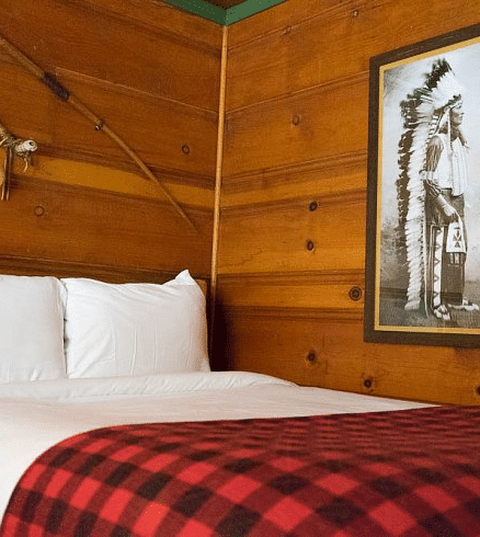 Overview of a Bed in a room at Shaver Lake Village Hotel with white pillows, red plaid blanket, wood paneled walls, framed art, and decorative sticks.
