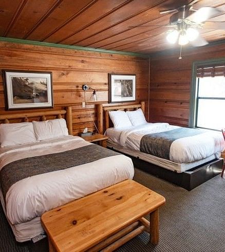 Overview of a Room at Shaver Lake Village Hotel with two twin beds, wood panelled walls, a wooden table, and red chairs by a window.