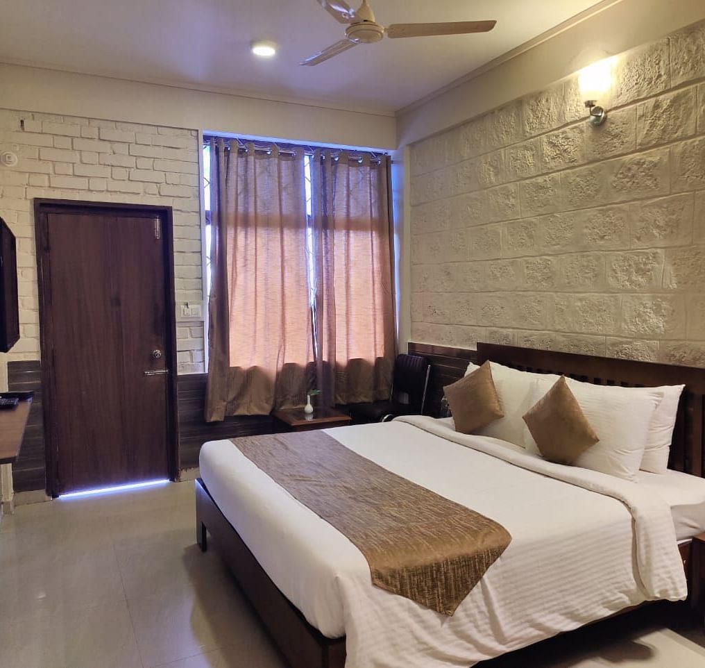 Bedroom with a huge bed, bedside table, windows with curtains, wooden door, and a TV | Standard Room | Woodstock Resorts, Coorg