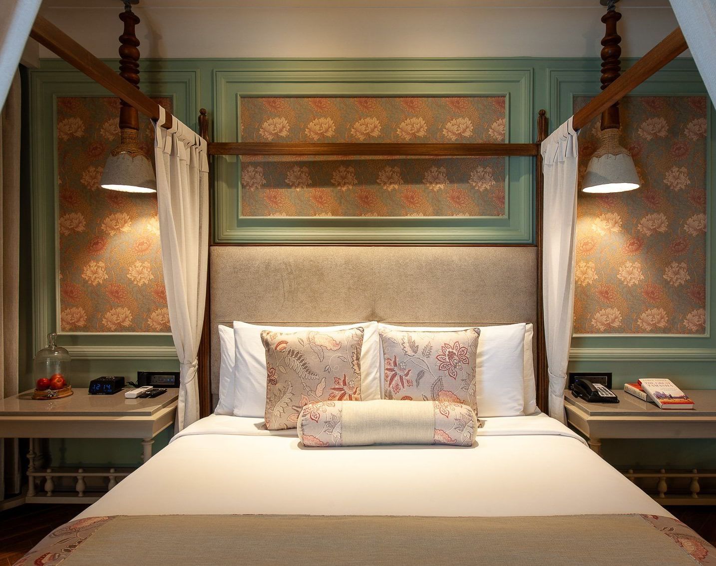 A close-up, front-facing view of the beautifully appointed four-poster bed in a suite at The Evren, Vagator. The bed is adorned with decorative pillows, and the background features elegant wallpaper and unique hanging lights.