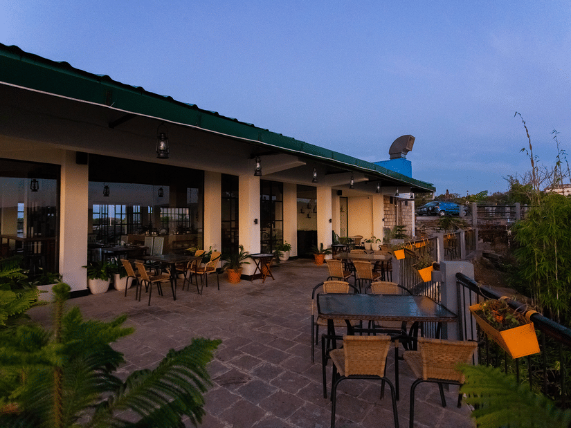 Open-air terrace cafe at Polo Resort Cherrapunji at dusk with wicker chairs, glass-top tables, hanging lanterns and orange planter boxes on the railing.