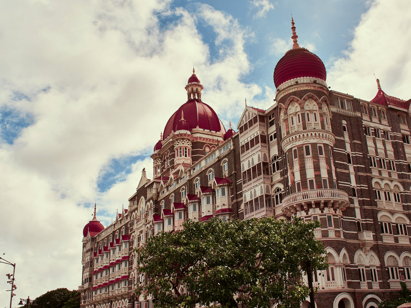The large Taj Hotel building with domes and arched windows, with trees in front and clouds in the background.