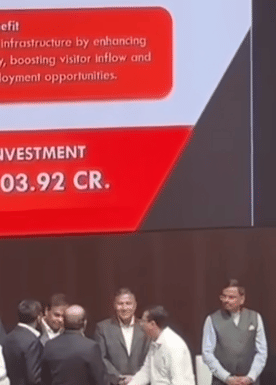 Men in formal attire stand on a stage in front of a red screen displaying tourism infrastructure benefit text and the figure Investment 103.92 CR.