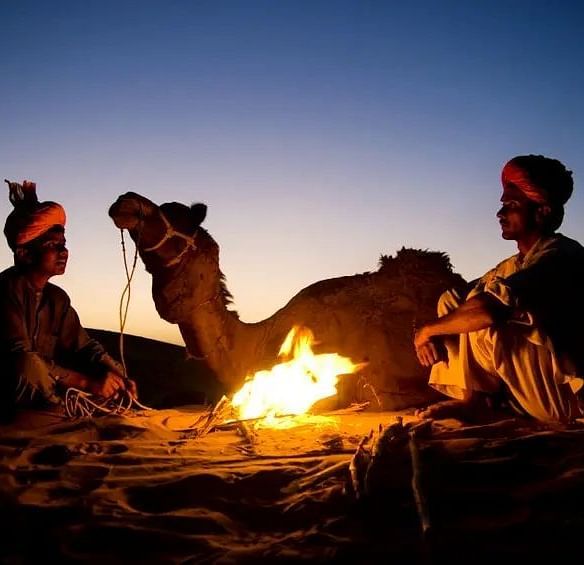 An image shot during dusk where two men are sitting near a bonfire and a camel is resting in the background