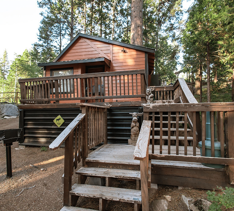 A Cabin at Shaver Lake Village Hotel with a wooden deck, stairs, and a grill, surrounded by tall pine trees.