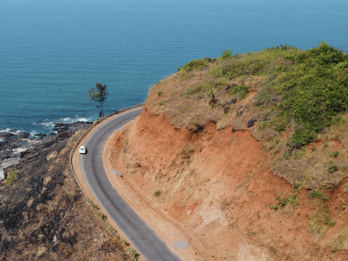 An aerial view showing a curved road winding along a coastline, positioned between a rocky shore by blue water and a grassy, reddish hillside inbackground.